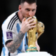 Messi kissing the FIFA World Cup trophy while holding it close in an Argentina jersey with the tournament patch visible on the sleeve