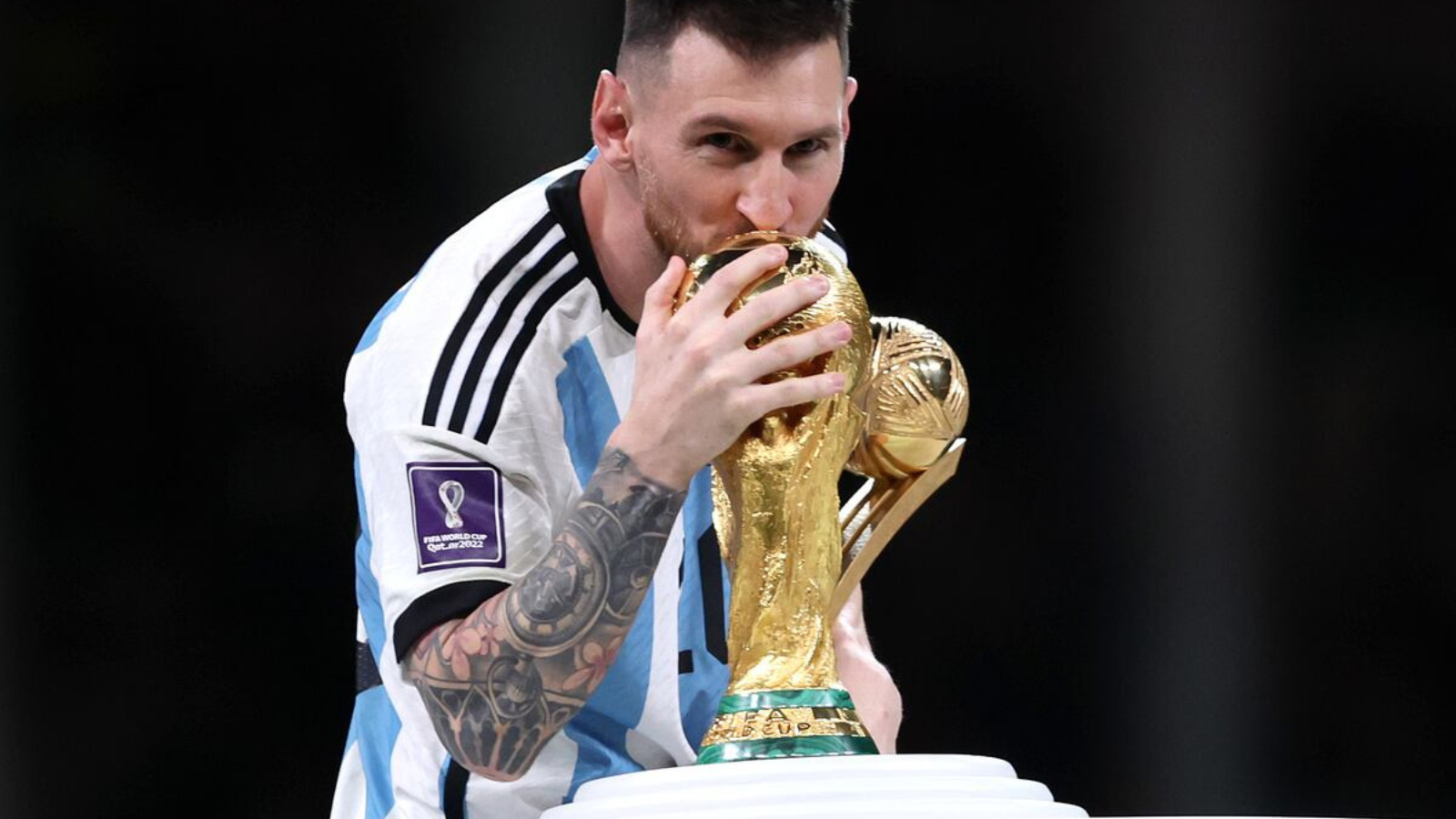 Messi kissing the FIFA World Cup trophy while holding it close in an Argentina jersey with the tournament patch visible on the sleeve