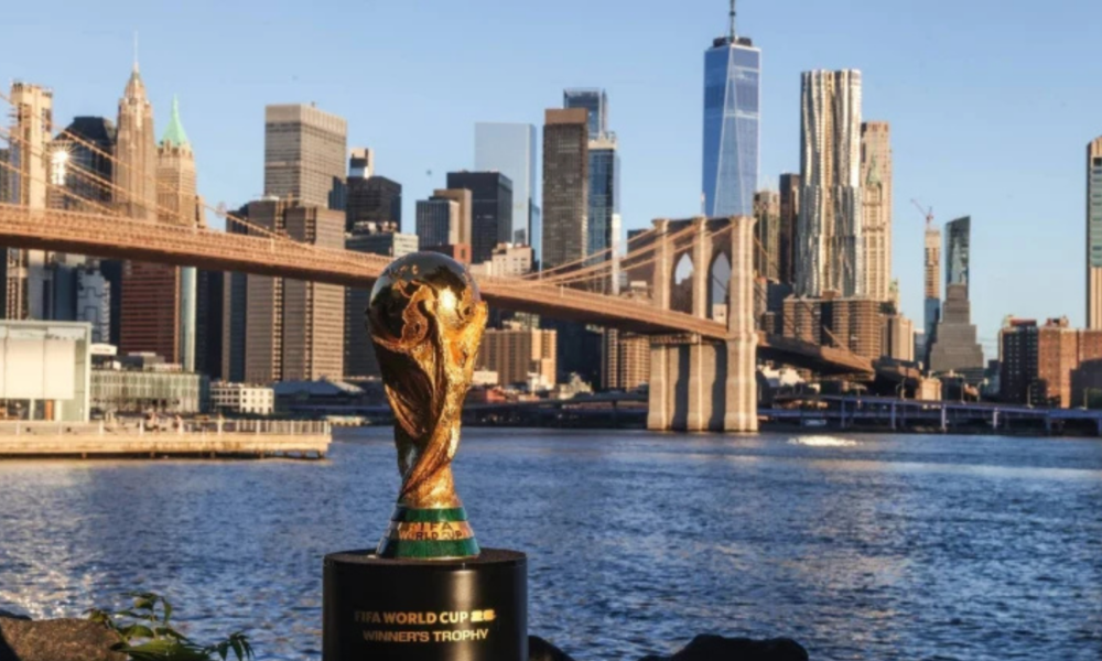 FIFA World Cup trophy on a pedestal by the river with the Brooklyn Bridge and Manhattan skyline in the background.