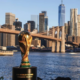 FIFA World Cup trophy on a pedestal by the river with the Brooklyn Bridge and Manhattan skyline in the background.