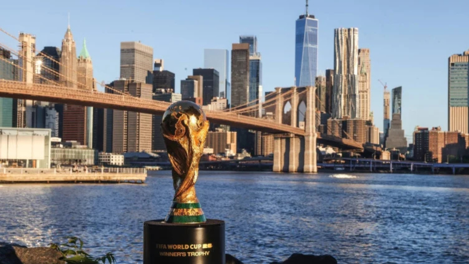 FIFA World Cup trophy on a pedestal by the river with the Brooklyn Bridge and Manhattan skyline in the background.