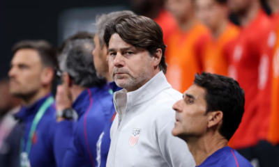 Man in a white USA jacket with crest, standing on the sidelines during a lineup.