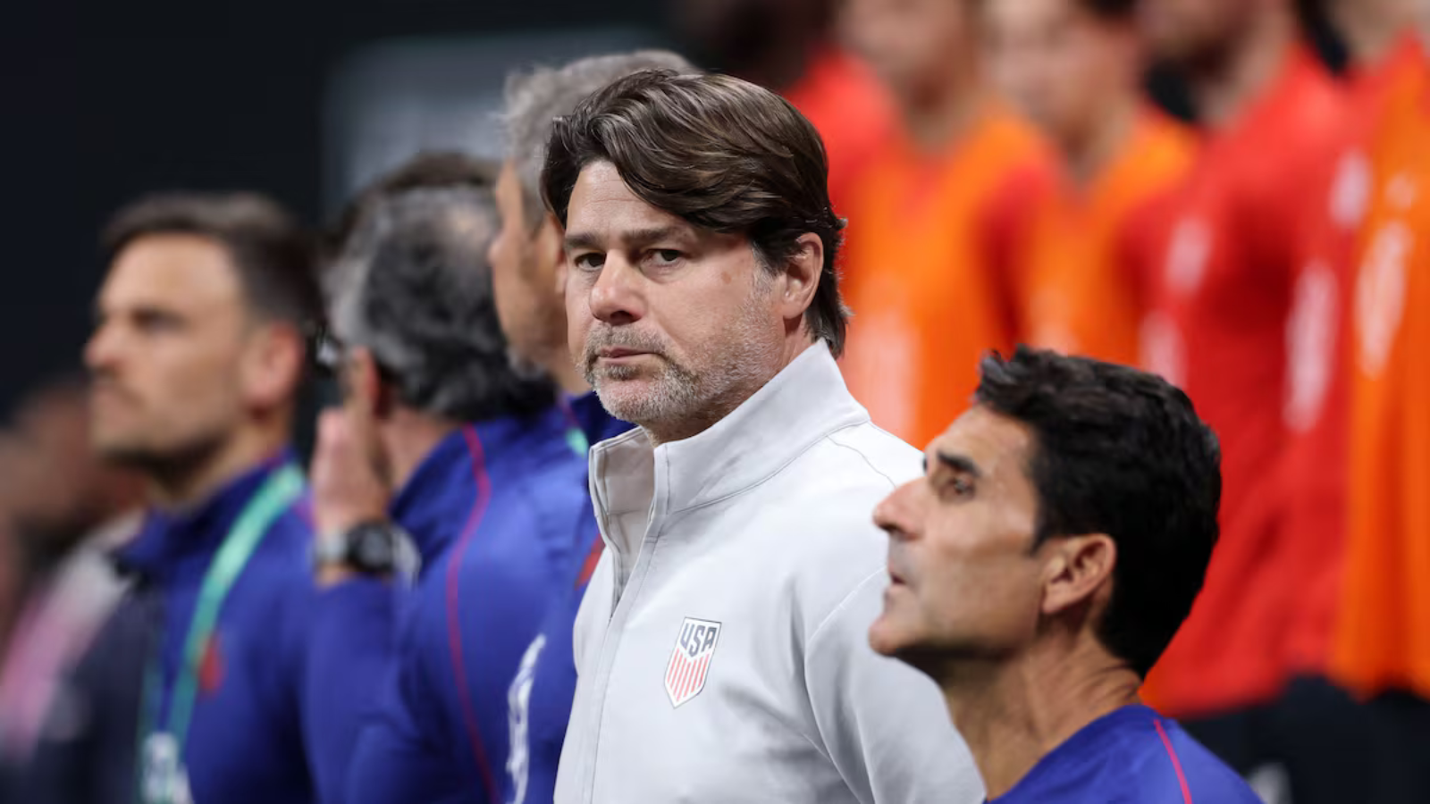 Man in a white USA jacket with crest, standing on the sidelines during a lineup.