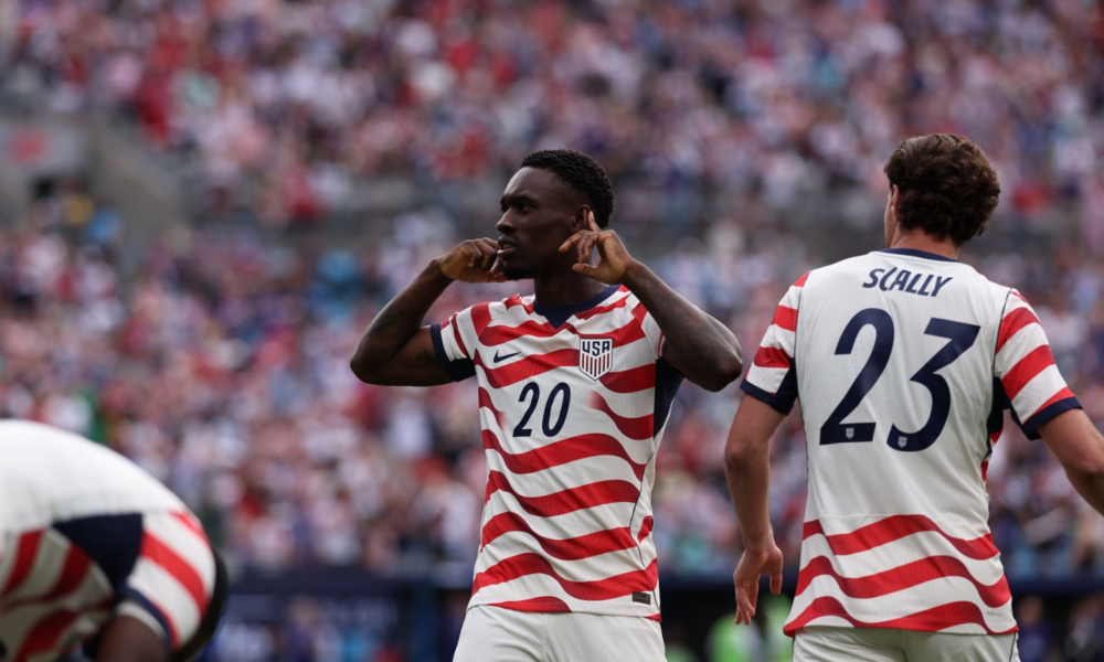 Two USA soccer players in red-and-white striped jerseys on the field; #20 gestures toward his ears while #23 (Scally) faces away amid a cheering crowd.