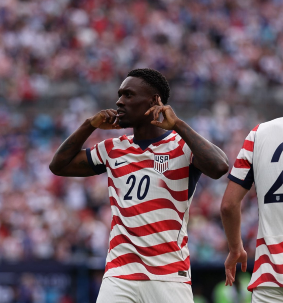 Two USA soccer players in red-and-white striped jerseys on the field; #20 gestures toward his ears while #23 (Scally) faces away amid a cheering crowd.