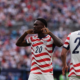 Two USA soccer players in red-and-white striped jerseys on the field; #20 gestures toward his ears while #23 (Scally) faces away amid a cheering crowd.