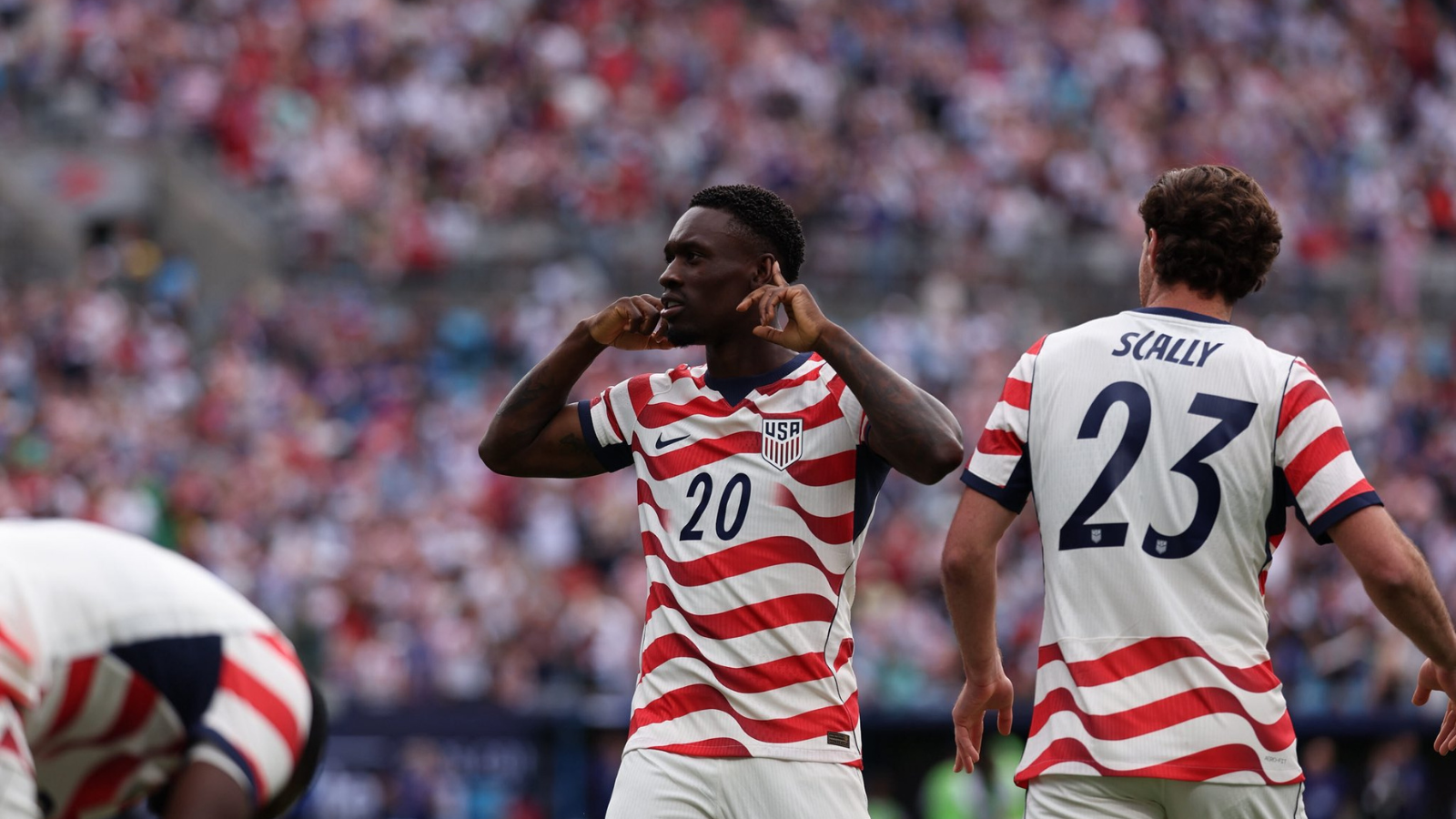 Two USA soccer players in red-and-white striped jerseys on the field; #20 gestures toward his ears while #23 (Scally) faces away amid a cheering crowd.