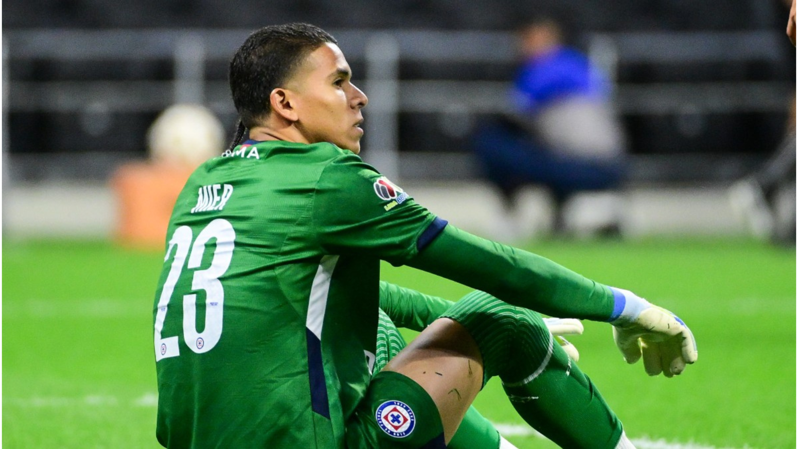 Soccer goalkeeper in a green kit with number 23 sits on the grass, knees up, during a match.