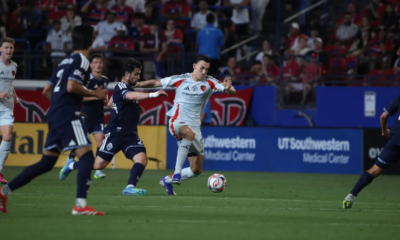 Player in white and red kit kicks the ball while defeated by two navy-clad defenders during a crowded match.