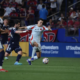 Player in white and red kit kicks the ball while defeated by two navy-clad defenders during a crowded match.
