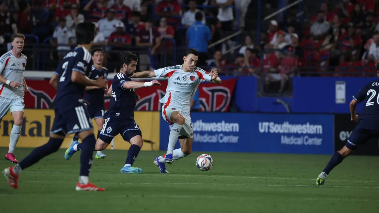 Player in white and red kit kicks the ball while defeated by two navy-clad defenders during a crowded match.