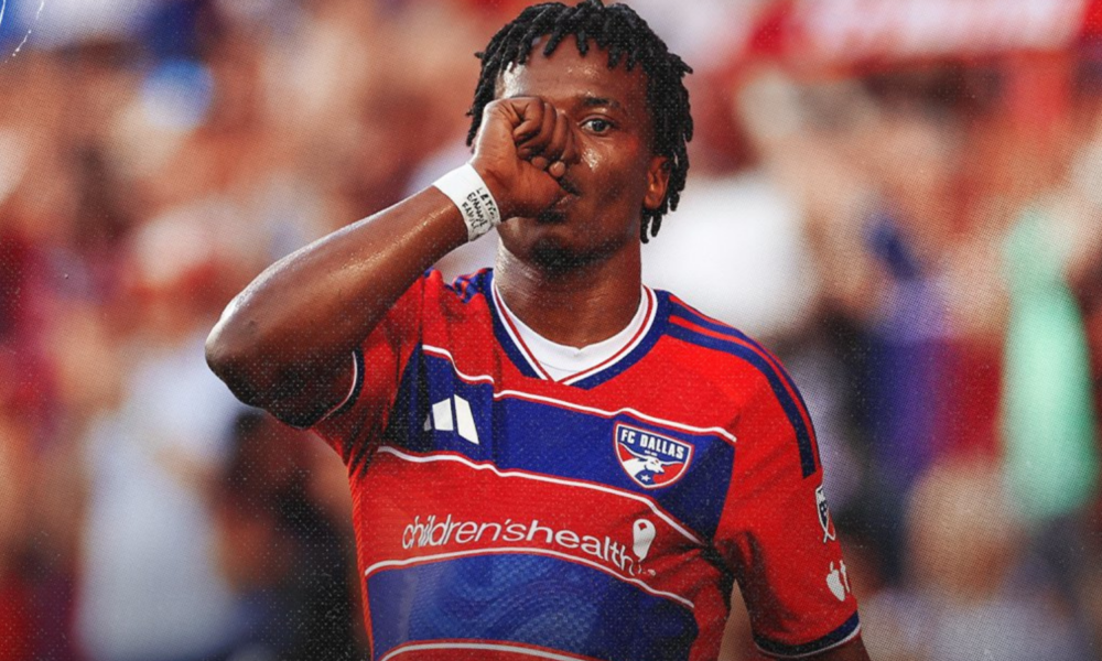 Soccer player in red FC Dallas jersey touching his mouth with his fist during a match, crowd blurred in the background.