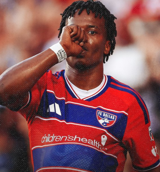 Soccer player in red FC Dallas jersey touching his mouth with his fist during a match, crowd blurred in the background.