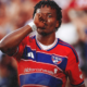 Soccer player in red FC Dallas jersey touching his mouth with his fist during a match, crowd blurred in the background.