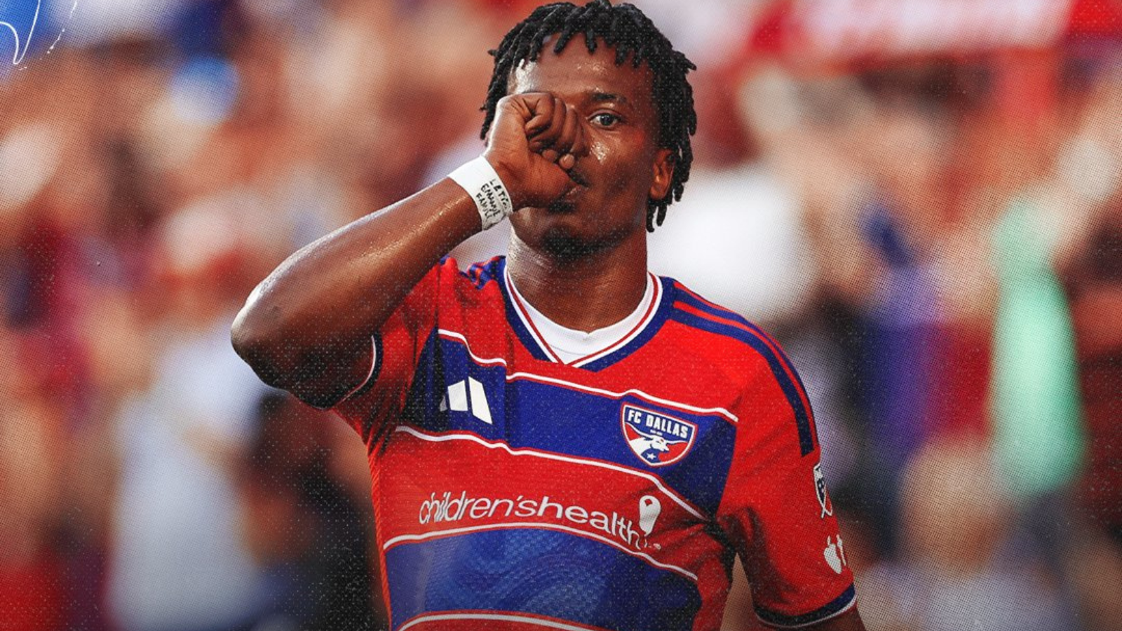 Soccer player in red FC Dallas jersey touching his mouth with his fist during a match, crowd blurred in the background.