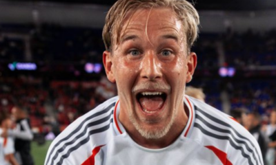 Close-up of a jubilant male footballer with wet hair, mouth open in celebration, wearing a white jersey with gray stripes on a stadium field