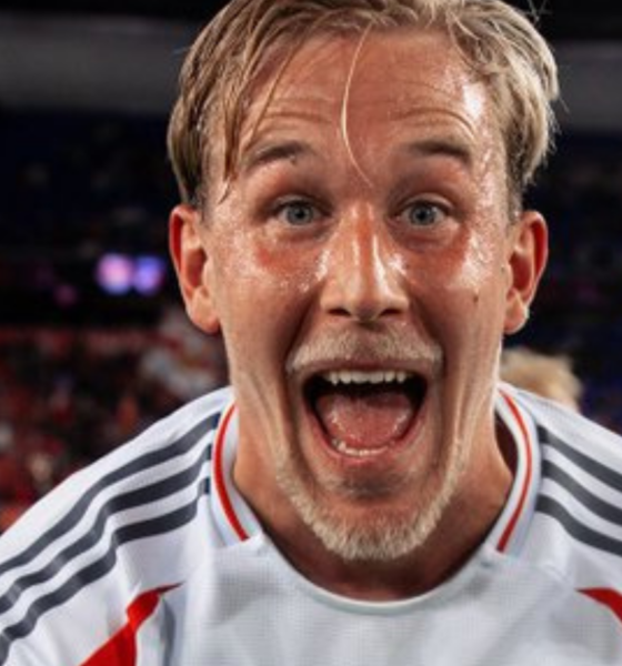 Close-up of a jubilant male footballer with wet hair, mouth open in celebration, wearing a white jersey with gray stripes on a stadium field