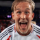 Close-up of a jubilant male footballer with wet hair, mouth open in celebration, wearing a white jersey with gray stripes on a stadium field