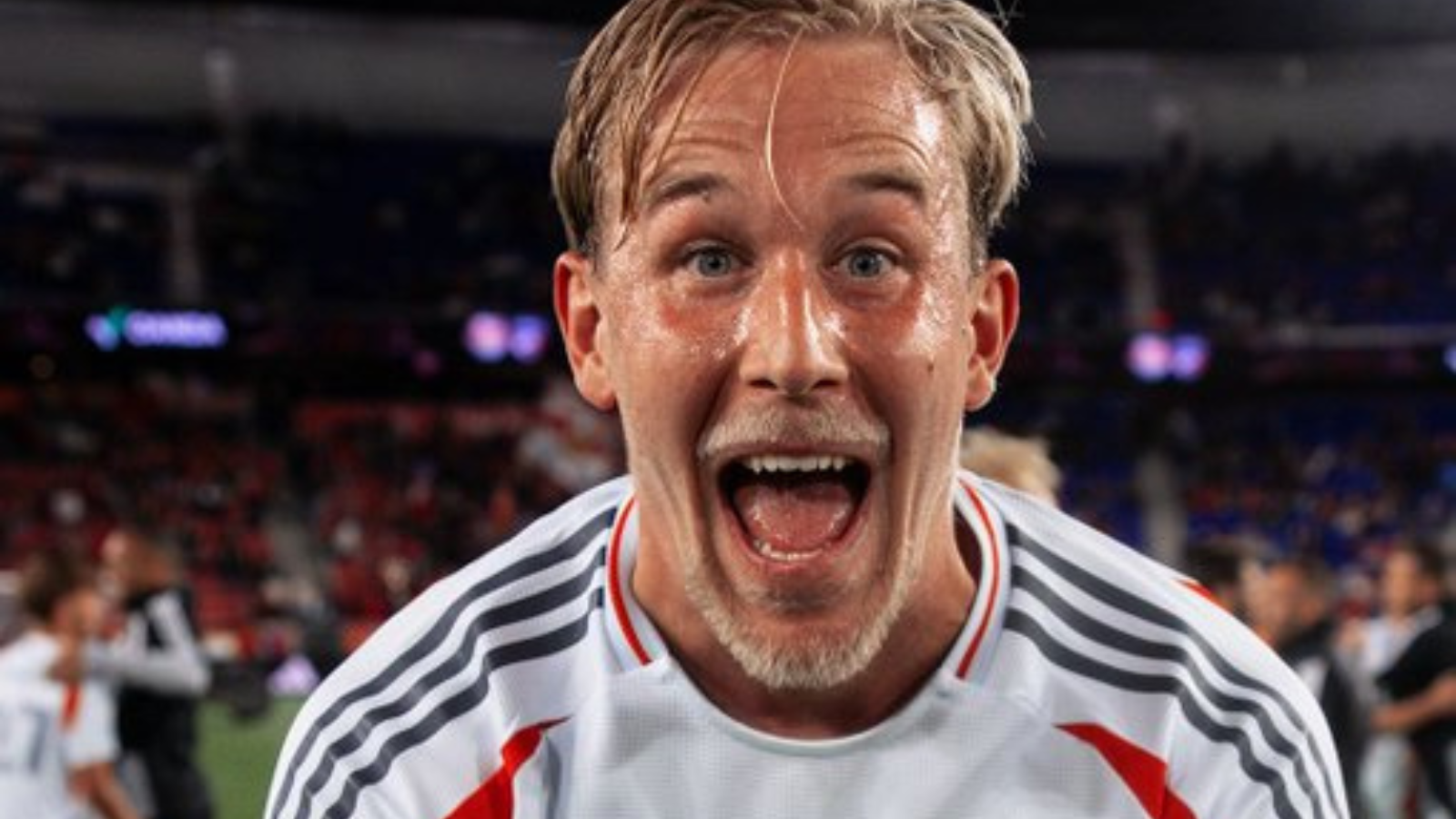 Close-up of a jubilant male footballer with wet hair, mouth open in celebration, wearing a white jersey with gray stripes on a stadium field