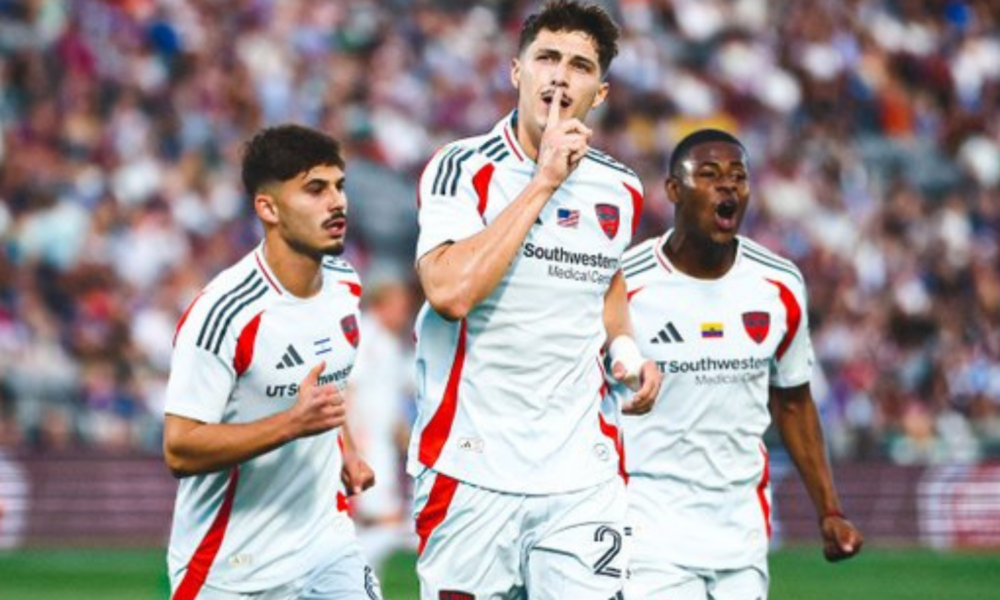 Three soccer players in white uniforms celebrate a goal on the field; the central player puts a finger to his lips in a hush gesture as teammates cheer.
