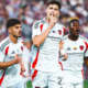 Three soccer players in white uniforms celebrate a goal on the field; the central player puts a finger to his lips in a hush gesture as teammates cheer.