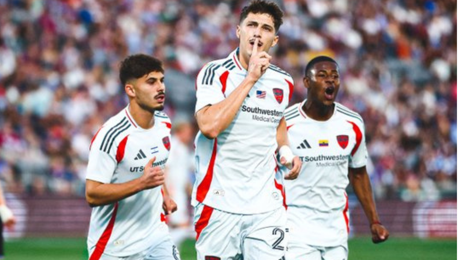 Three soccer players in white uniforms celebrate a goal on the field; the central player puts a finger to his lips in a hush gesture as teammates cheer.