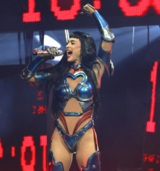 Female performer in blue metallic costume singing into a microphone on stage with red LED screens behind him/her.