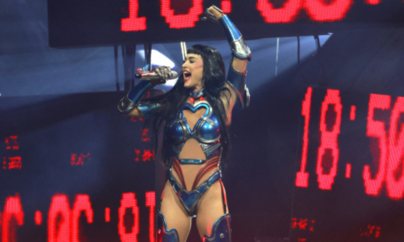 Female performer in blue metallic costume singing into a microphone on stage with red LED screens behind him/her.