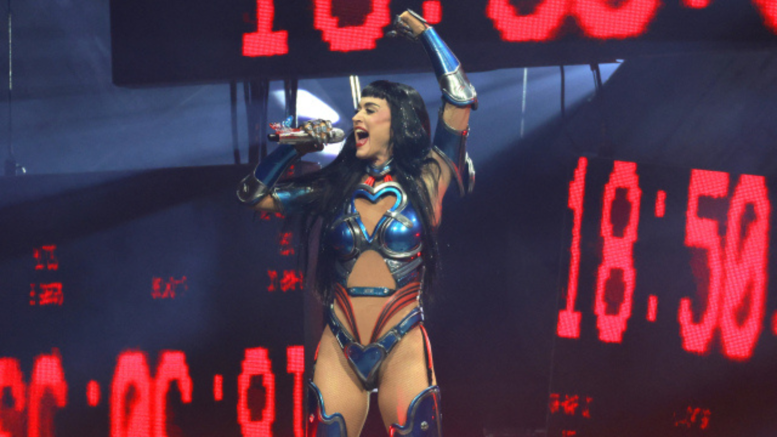Female performer in blue metallic costume singing into a microphone on stage with red LED screens behind him/her.
