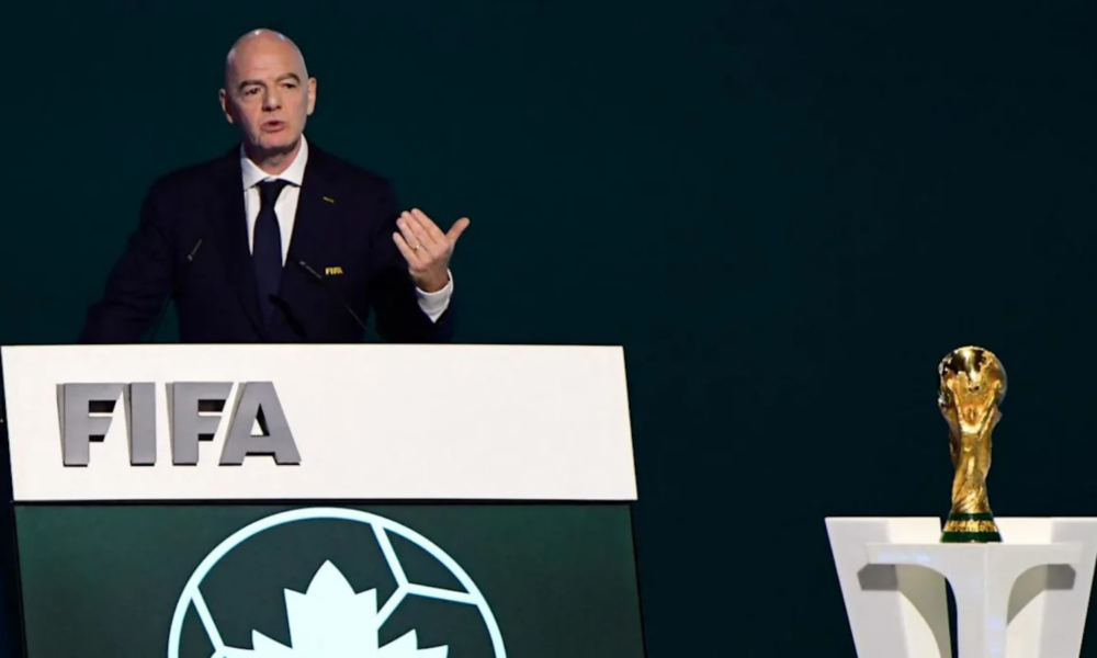 Man delivering a speech at a FIFA podium, with a World Cup trophy on a pedestal to the right.