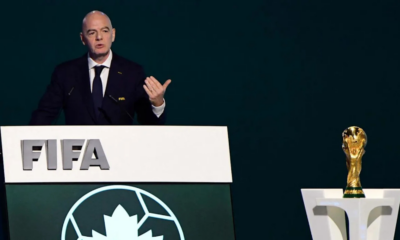 Man delivering a speech at a FIFA podium, with a World Cup trophy on a pedestal to the right.
