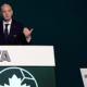 Man delivering a speech at a FIFA podium, with a World Cup trophy on a pedestal to the right.