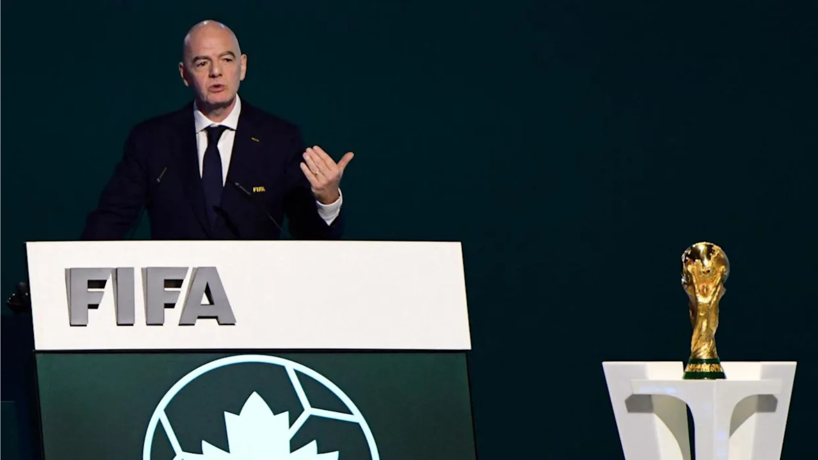 Man delivering a speech at a FIFA podium, with a World Cup trophy on a pedestal to the right.
