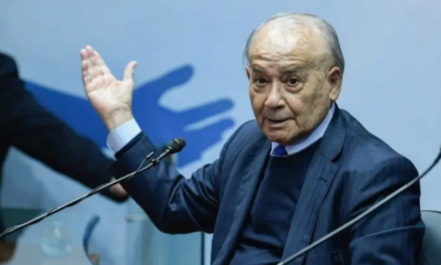Older man in a dark suit raises his left hand while speaking into a microphone on a panel stage.
