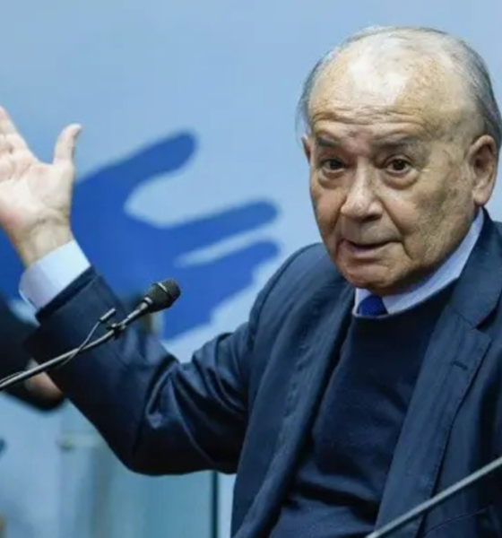Older man in a dark suit raises his left hand while speaking into a microphone on a panel stage.