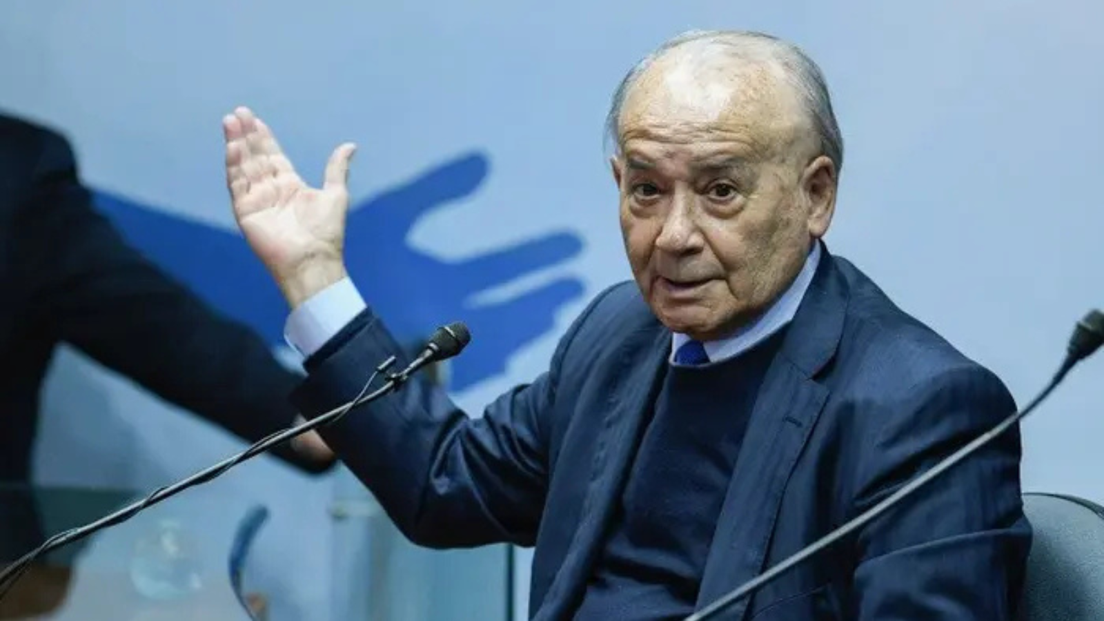 Older man in a dark suit raises his left hand while speaking into a microphone on a panel stage.