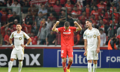 Soccer player in red raises arms in celebration between two white-clad teammates on a filled stadium field with fans behind him (celebration scene).