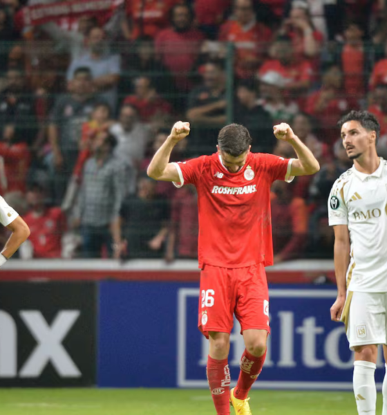 Soccer player in red raises arms in celebration between two white-clad teammates on a filled stadium field with fans behind him (celebration scene).