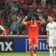 Soccer player in red raises arms in celebration between two white-clad teammates on a filled stadium field with fans behind him (celebration scene).