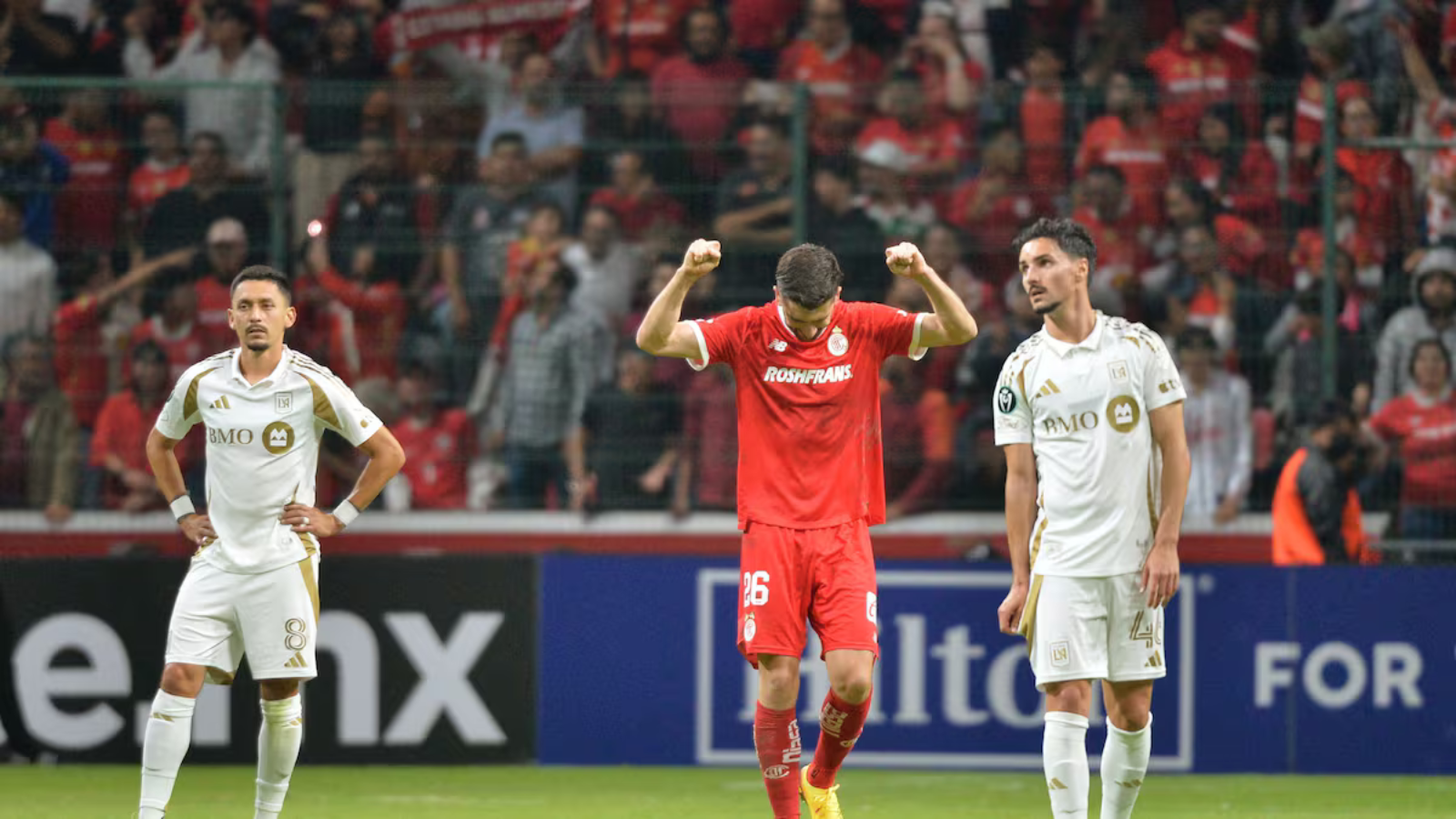 Soccer player in red raises arms in celebration between two white-clad teammates on a filled stadium field with fans behind him (celebration scene).