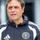 Male coach in a black-and-white training jacket with Miami FC crest, looking to the left outside.