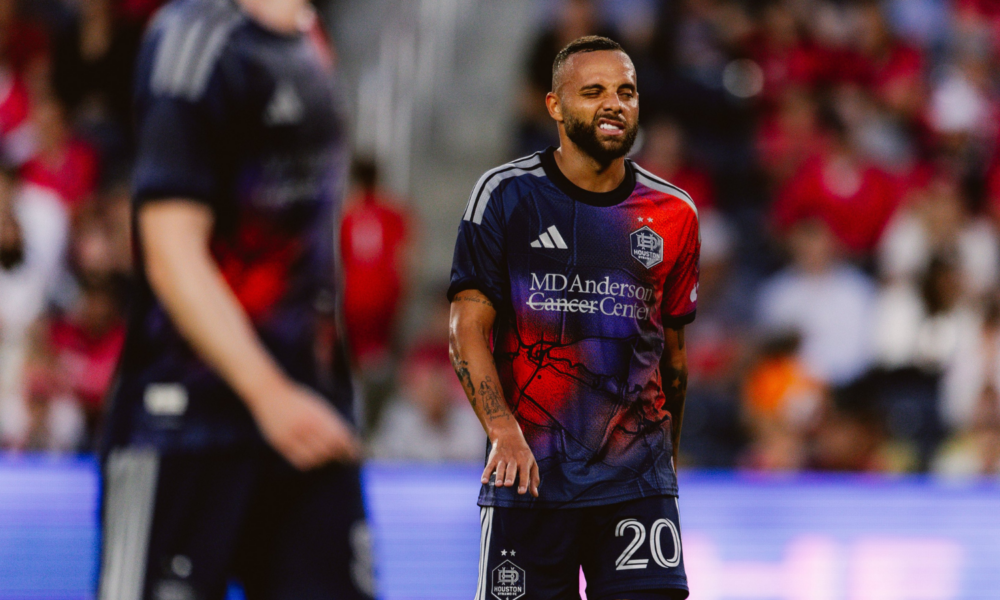 Soccer player in a blue-red Houston Dynamo jersey with number 20, grimacing on the field with a blurred crowd behind him, during a match.
