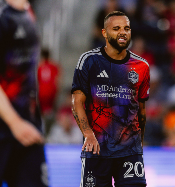 Soccer player in a blue-red Houston Dynamo jersey with number 20, grimacing on the field with a blurred crowd behind him, during a match.
