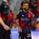 Soccer player in a blue-red Houston Dynamo jersey with number 20, grimacing on the field with a blurred crowd behind him, during a match.