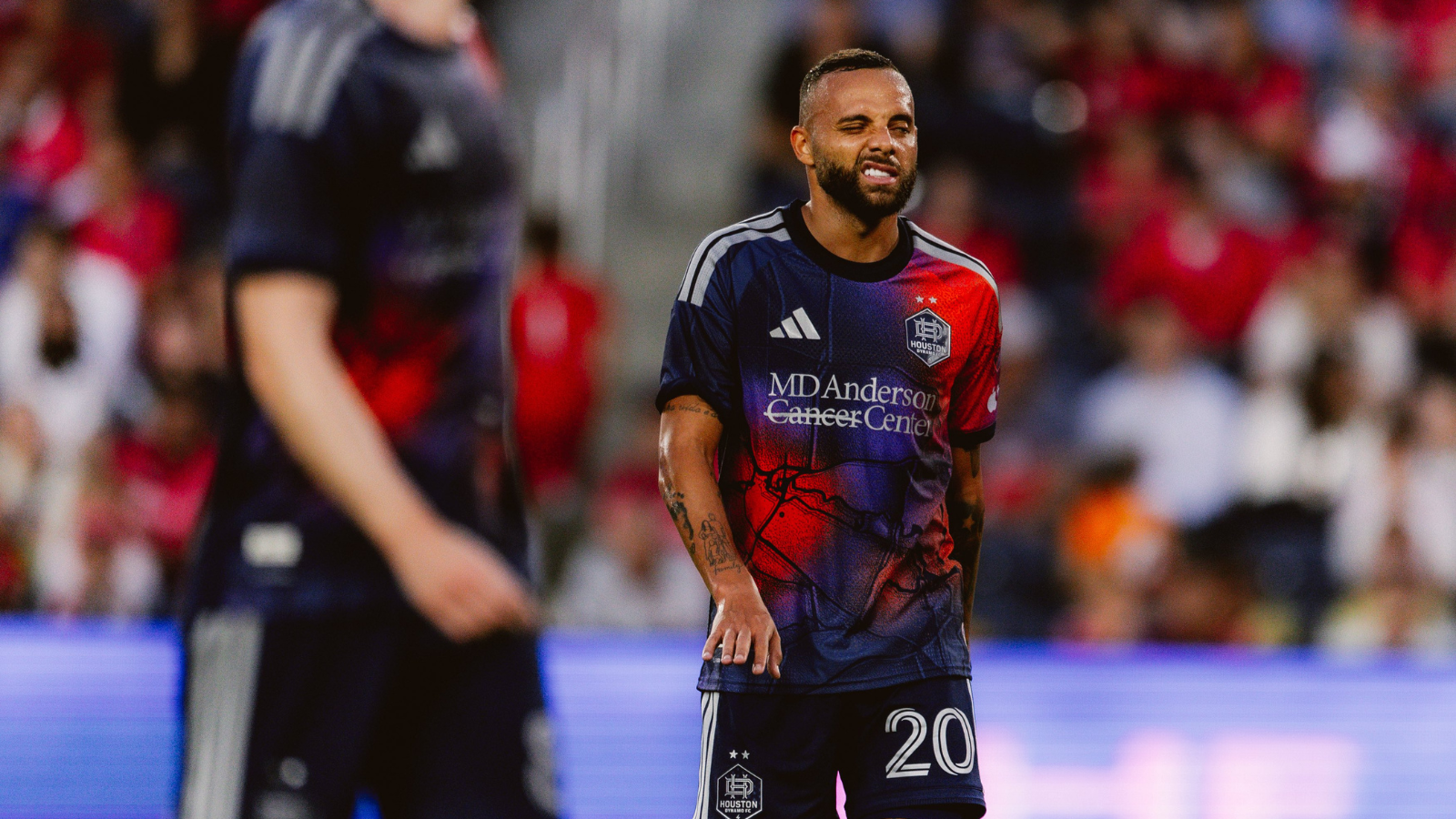 Soccer player in a blue-red Houston Dynamo jersey with number 20, grimacing on the field with a blurred crowd behind him, during a match.