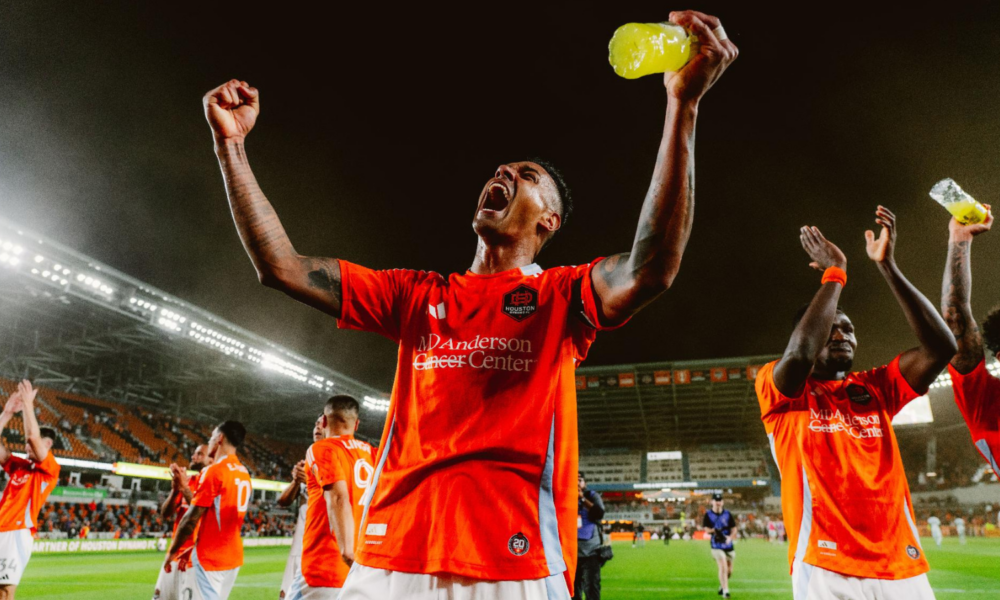 Soccer players in red jerseys celebrate on the pitch at night, central player with arms raised and yellow drink bottle raised high, teammates around him.