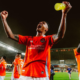 Soccer players in red jerseys celebrate on the pitch at night, central player with arms raised and yellow drink bottle raised high, teammates around him.