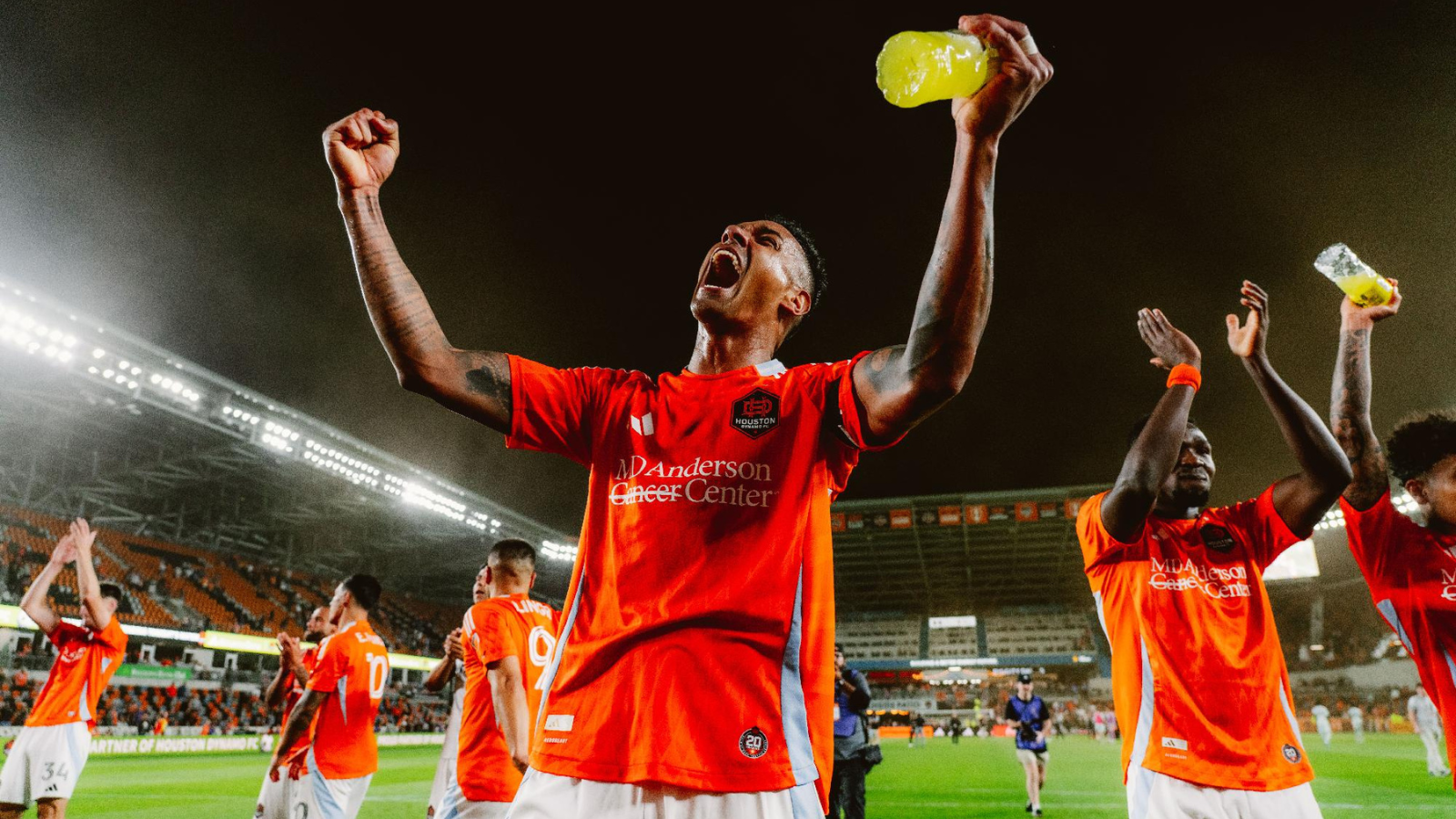 Soccer players in red jerseys celebrate on the pitch at night, central player with arms raised and yellow drink bottle raised high, teammates around him.