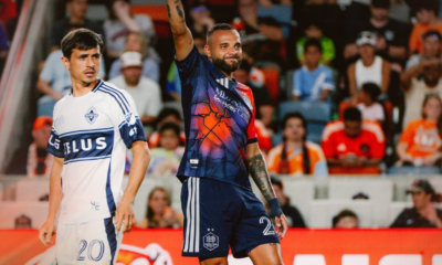 Two soccer players on the field during a match; one in a white jersey with number 20, the other in a dark blue kit raising his arm amid a cheering crowd.