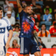 Two soccer players on the field during a match; one in a white jersey with number 20, the other in a dark blue kit raising his arm amid a cheering crowd.